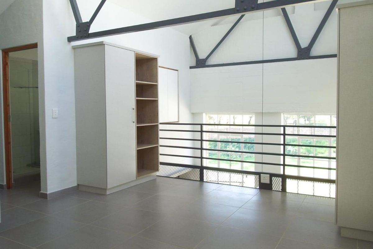Spacious loft-style bedroom featuring exposed steel roof trusses, tiled flooring, built-in cabinetry, and a metal balustrade overlooking the lower level.