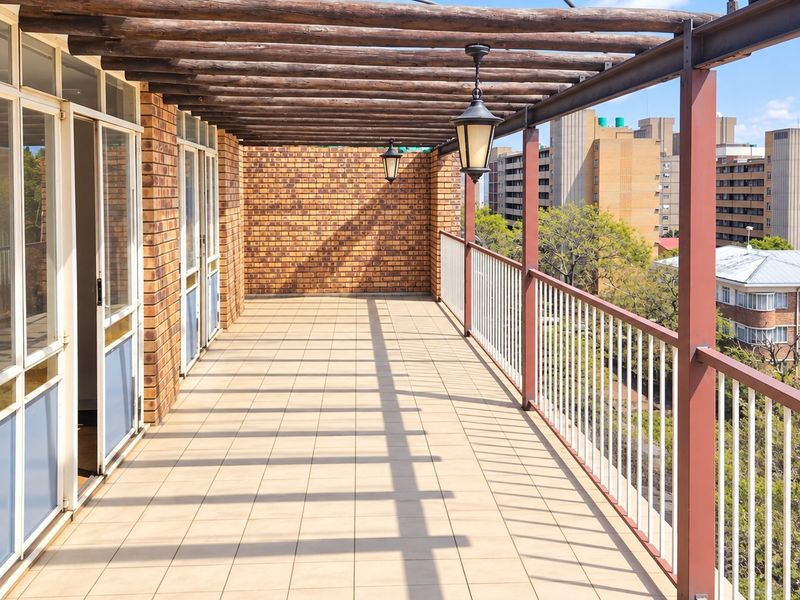 Spacious covered balcony featuring a timber pergola, tiled flooring, and a metal balustrade, offering an open-air space with views of the surrounding apartment complex.