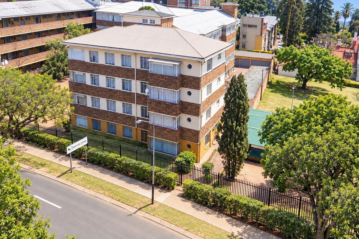 Aerial view of a multi-story brick apartment building featuring a pitched roof, perimeter fencing, and surrounding landscaped grounds.
