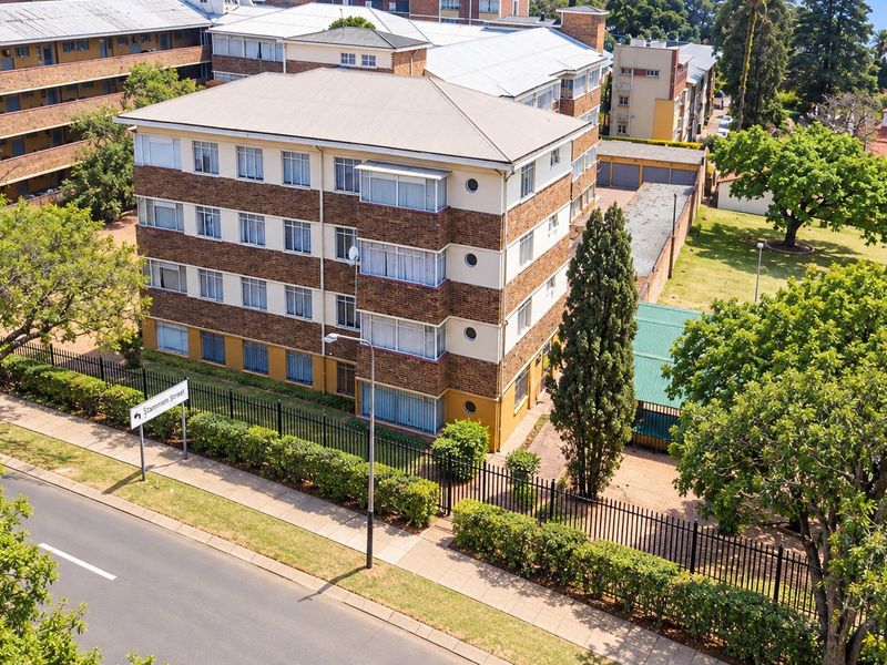 Aerial view of a multi-story brick apartment building featuring a pitched roof, perimeter fencing, and surrounding landscaped grounds.