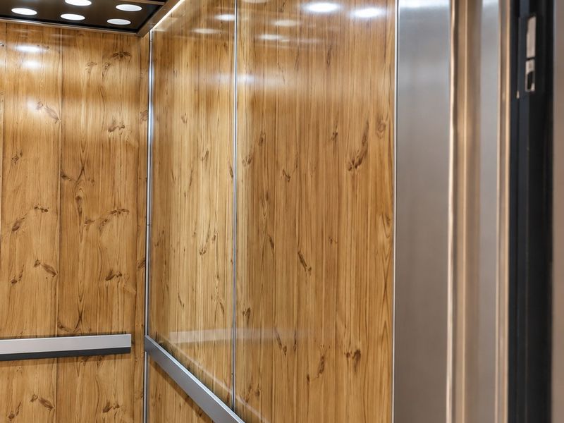 Interior view of a modern elevator cabin featuring wood-look wall paneling, recessed ceiling lighting, and stainless steel trim.