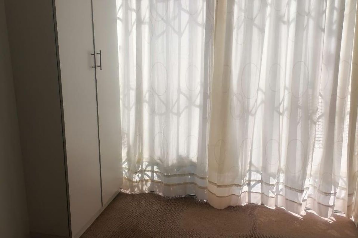 A vacant bedroom featuring built-in wardrobe storage and carpeted flooring, with natural light filtering through window curtains.