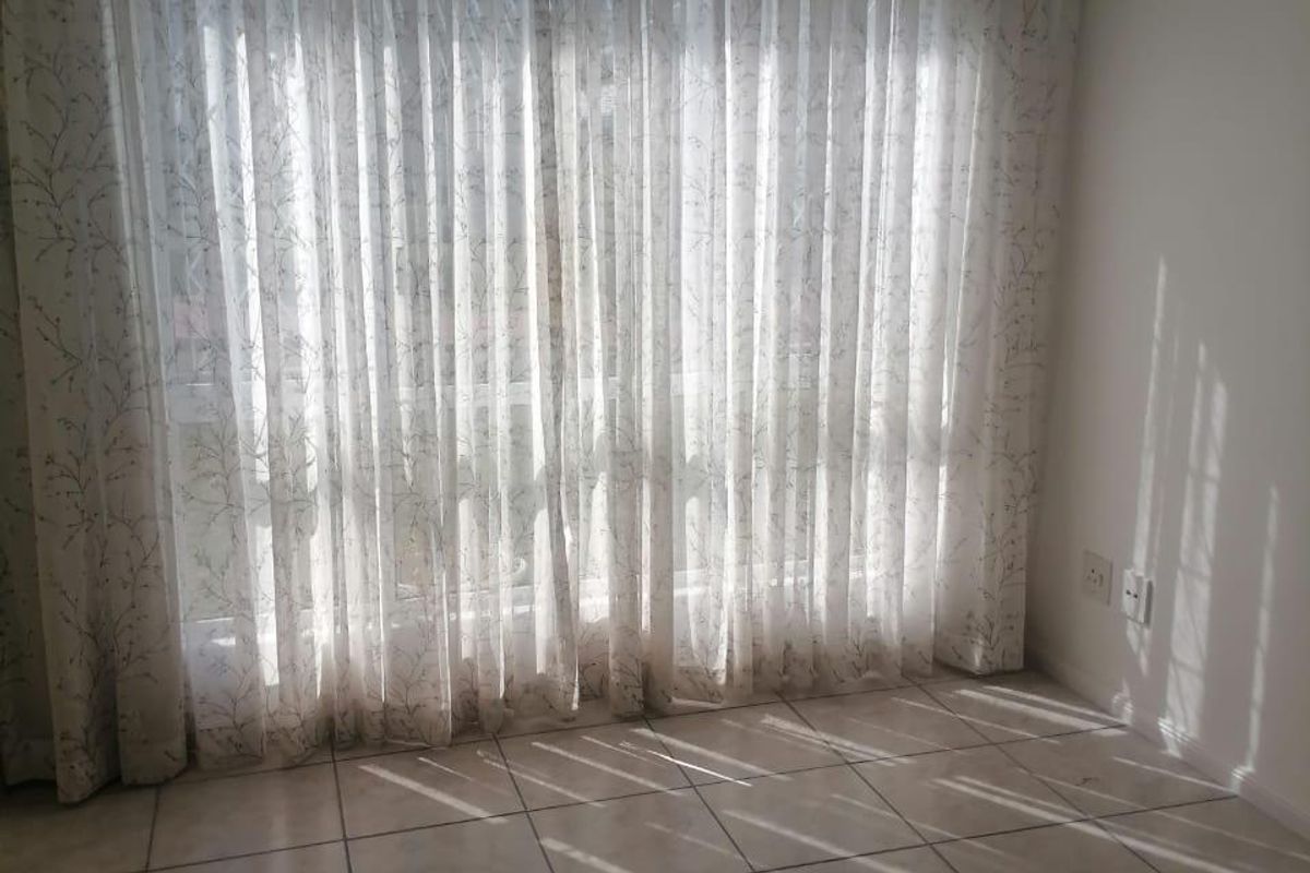 An empty living area featuring neutral tiled flooring, a ceiling fan, and large windows dressed with sheer curtains allowing natural light.