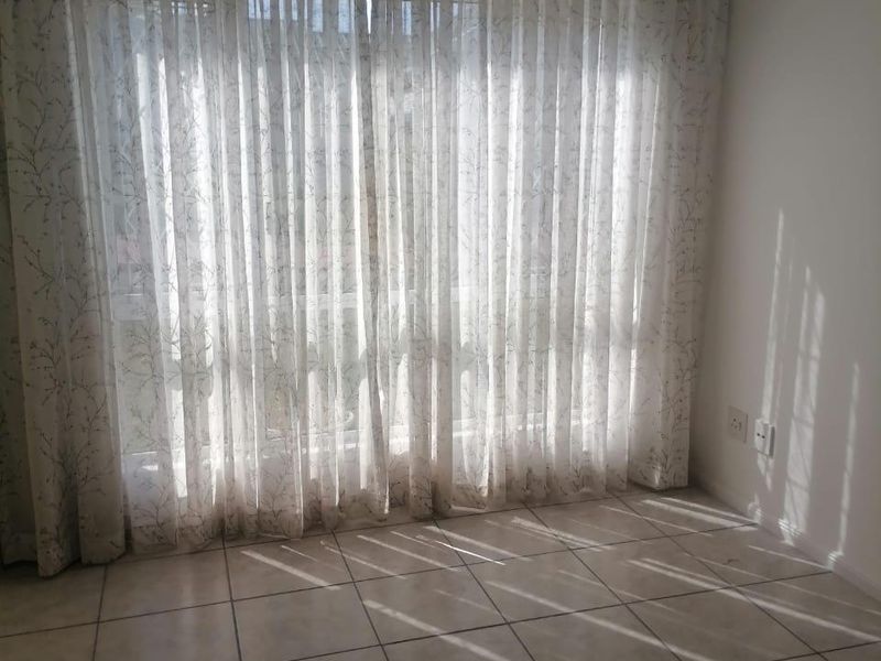 An empty living area featuring neutral tiled flooring, a ceiling fan, and large windows dressed with sheer curtains allowing natural light.
