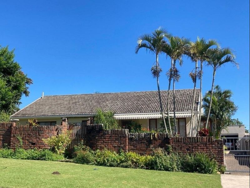 Front view of a single-story house featuring a brick boundary wall, tiled roof, and established garden with palm trees.