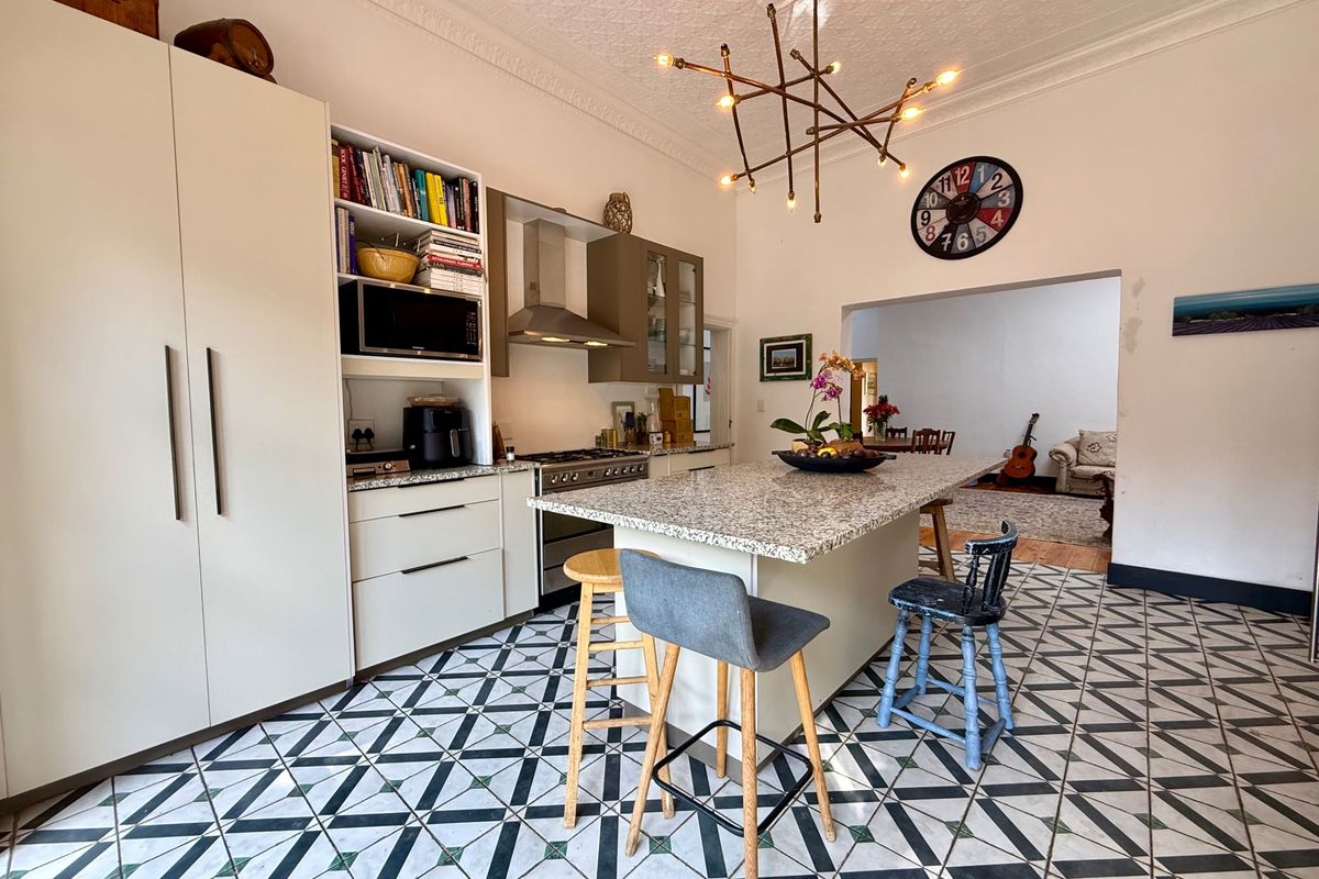 A well-appointed kitchen featuring a central island with granite countertops, patterned floor tiles, and decorative ceiling molding, opening into an adjacent living space.