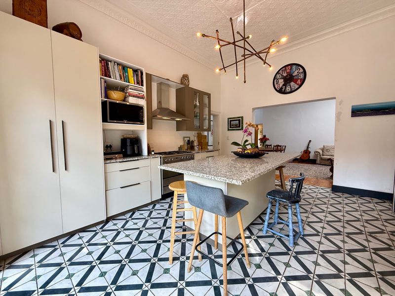 A well-appointed kitchen featuring a central island with granite countertops, patterned floor tiles, and decorative ceiling molding, opening into an adjacent living space.
