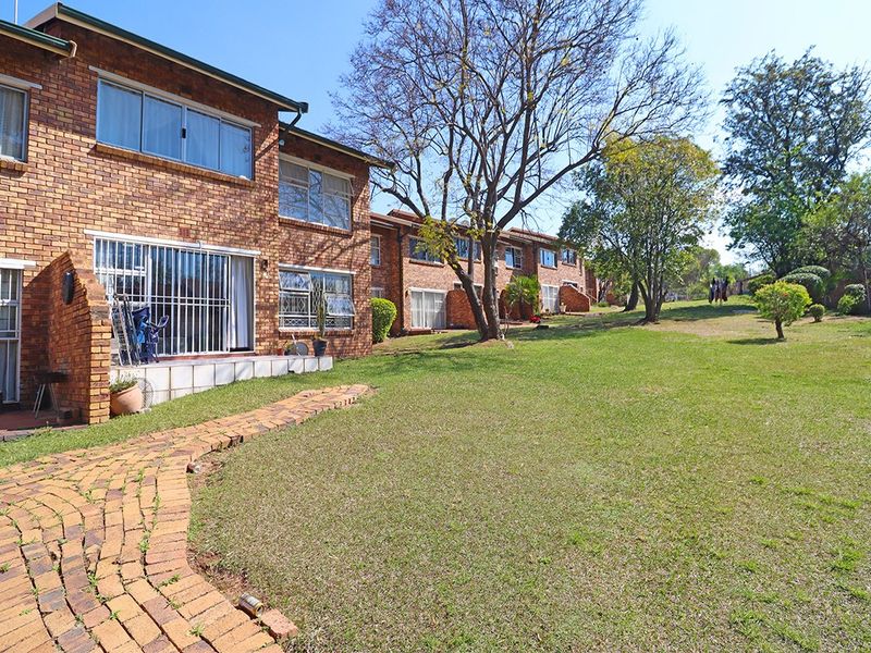 Rear view of a brick apartment building featuring a paved walkway, established lawn, and mature trees in a communal garden setting.