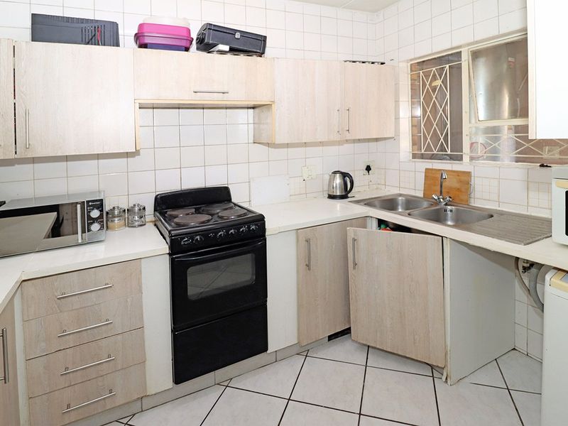 A functional kitchen featuring light wood-finish cabinetry, tiled backsplash, double stainless steel sink, and tiled flooring, requiring minor maintenance to cabinet hardware.