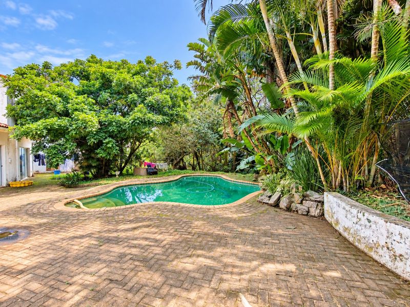 Outdoor pool area featuring a kidney-shaped swimming pool surrounded by brick paving and established garden landscaping.