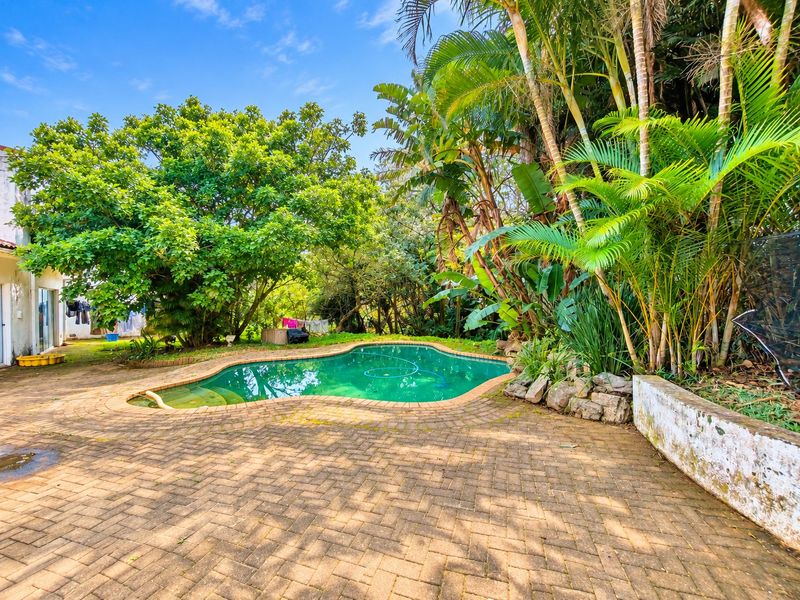 A private outdoor pool area featuring a kidney-shaped swimming pool surrounded by established tropical landscaping and brick paved patio space.