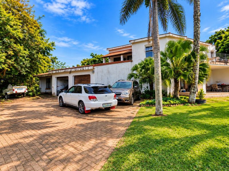 Front elevation of a two-story house featuring a paved driveway, double garage, and established garden with mature palm trees.