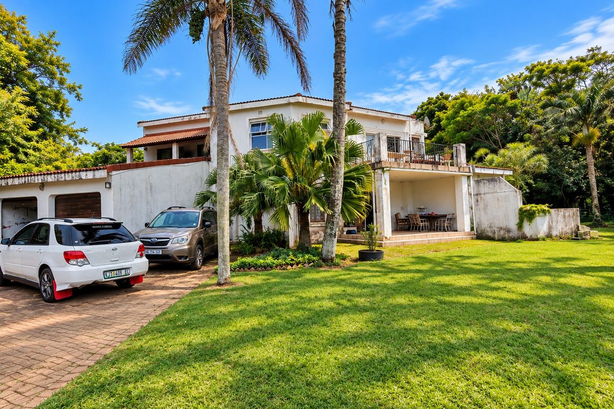 Rear view of a two-story house featuring a paved driveway, double garage, covered ground-floor patio, and an upper-level balcony with mature garden surroundings.