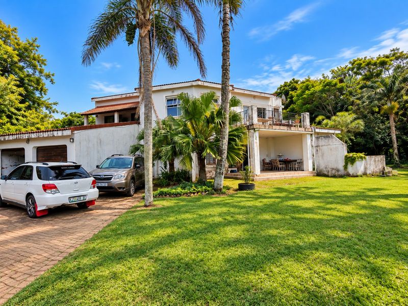 Rear view of a two-story house featuring a paved driveway, double garage, covered ground-floor patio, and an upper-level balcony with mature garden surroundings.