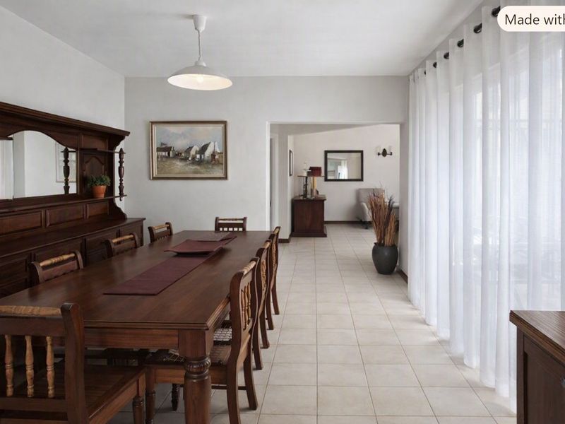 A spacious dining room featuring neutral tiled flooring, pendant lighting, and large windows with floor-to-ceiling curtains, leading into an adjacent living area.