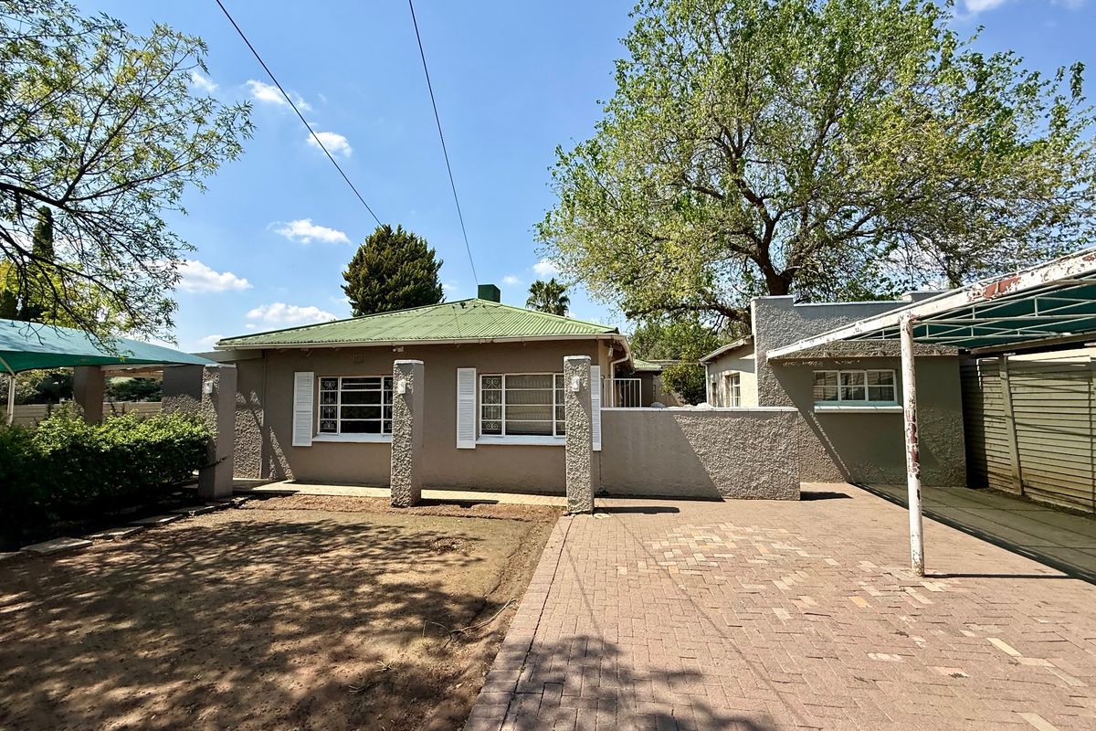 Rear view of the property featuring a paved driveway, carport, and established garden area with a corrugated metal roof.