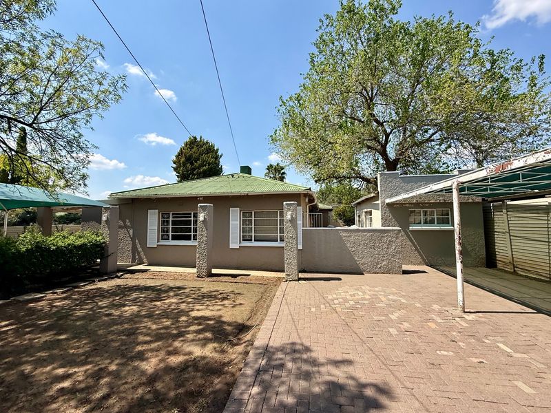 Rear view of the property featuring a paved driveway, carport, and established garden area with a corrugated metal roof.