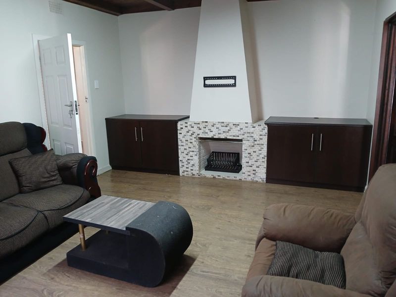 A living room featuring a central fireplace with mosaic tiling, built-in dark wood cabinetry, laminate flooring, and exposed timber ceiling beams.