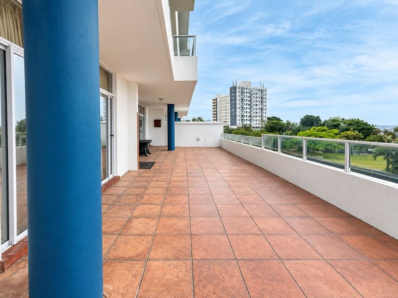 Spacious covered balcony featuring tiled flooring, glass balustrades, and structural columns, offering an open outdoor area with views.