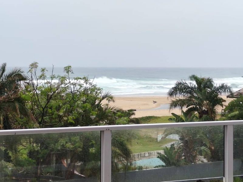 A scenic view of the ocean and beach from an elevated balcony featuring a modern glass balustrade.