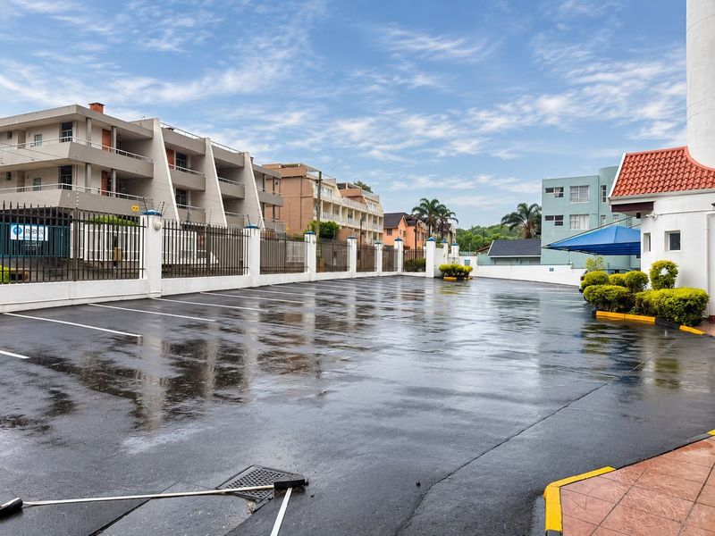 A paved outdoor parking area featuring perimeter security fencing and a shade sail structure within an apartment complex setting.