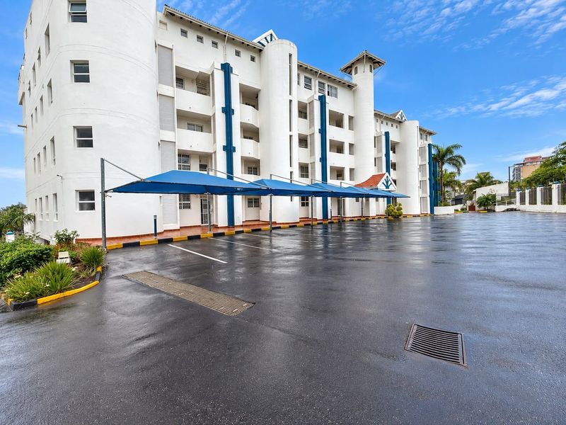 Exterior view of a multi-story apartment complex featuring a paved parking area with covered shade cloth carports and integrated drainage systems.