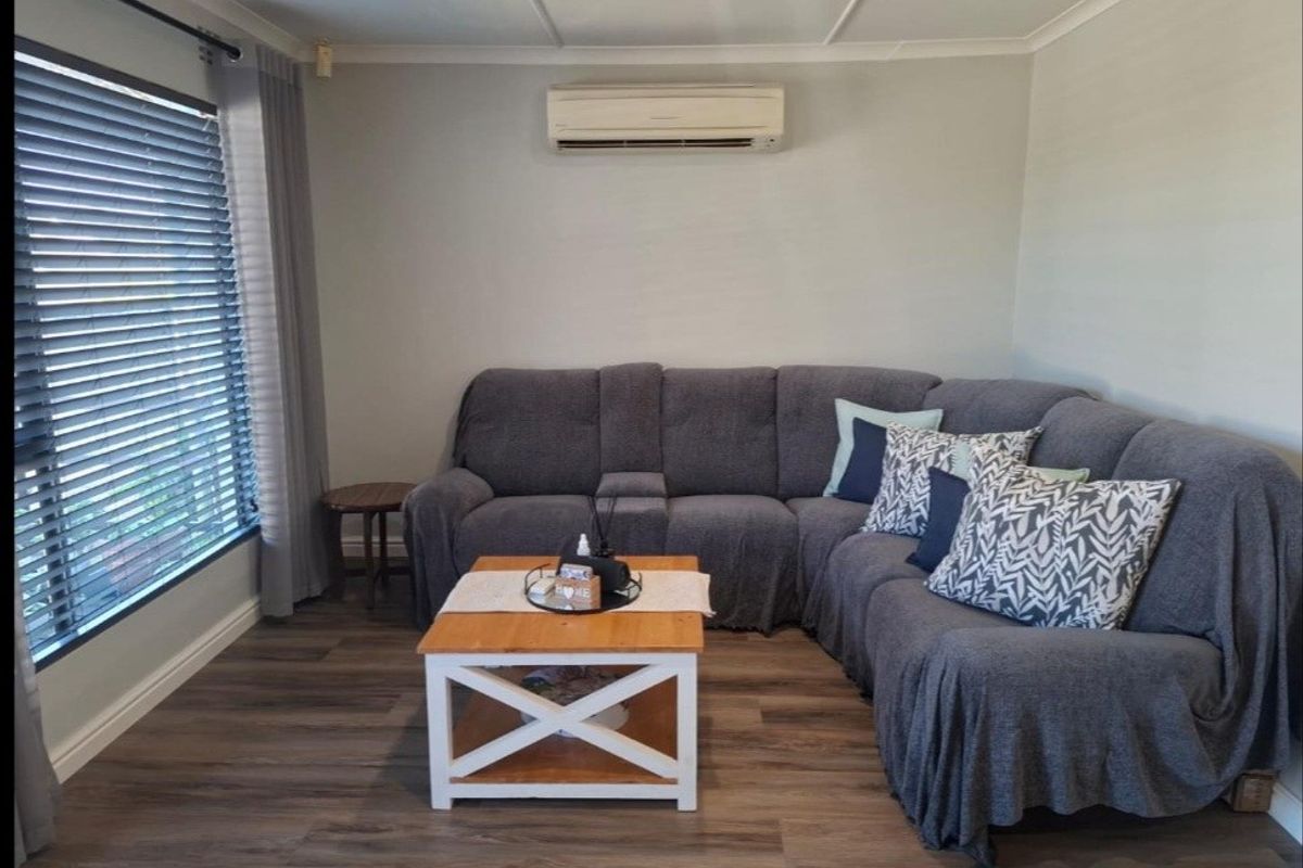 A comfortable living room featuring laminate flooring, a wall-mounted split-system air conditioner, and large windows with venetian blinds.