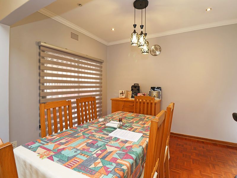 A well-maintained dining room featuring warm parquet flooring, recessed downlighting, a decorative pendant light, and modern roller blinds.