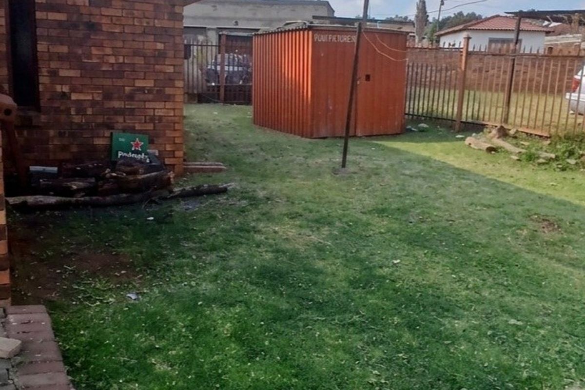 A view of the rear yard featuring a brick exterior wall, metal perimeter fencing, and a grass lawn area.