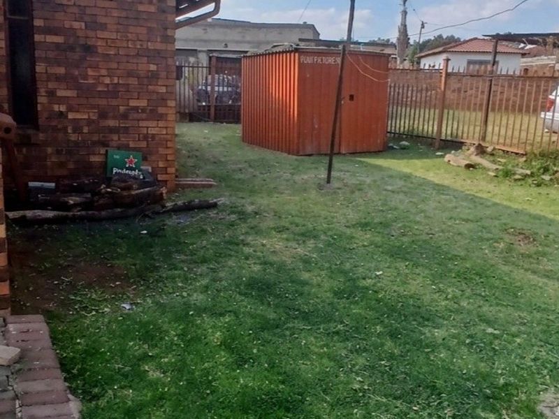 A view of the rear yard featuring a brick exterior wall, metal perimeter fencing, and a grass lawn area.