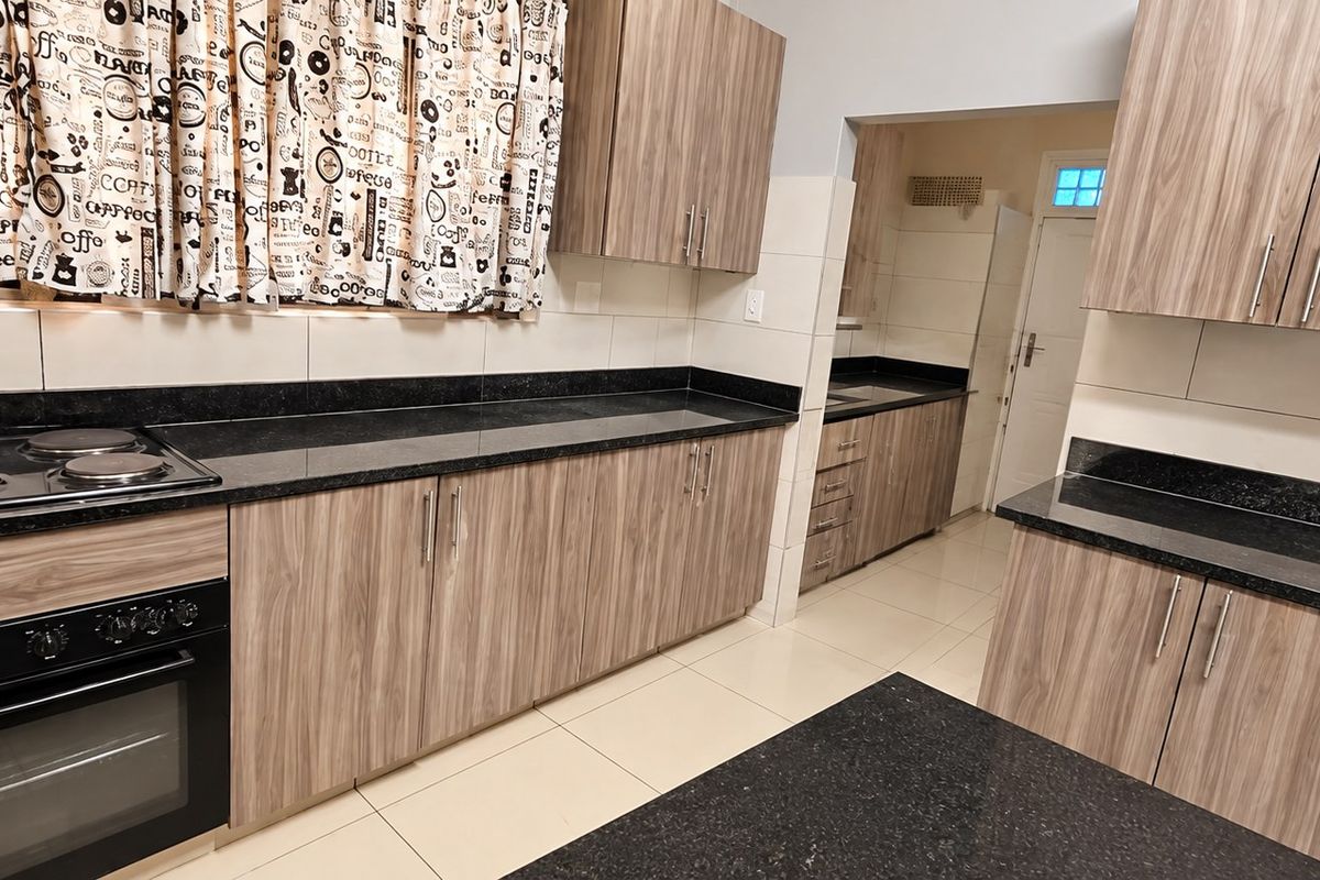 A modern kitchen featuring wood-grain cabinetry, black granite countertops, an integrated electric hob and oven, and neutral tiled flooring.