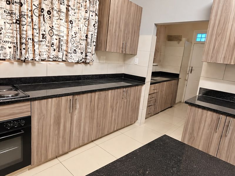 A modern kitchen featuring wood-grain cabinetry, black granite countertops, an integrated electric hob and oven, and neutral tiled flooring.