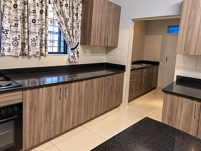 Modern kitchen featuring wood-grain cabinetry, dark granite countertops, built-in oven, electric hob, and a separate scullery area with additional storage and sink.