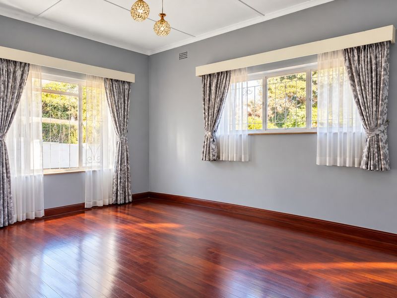 A bright and spacious living room featuring polished hardwood flooring, large windows with security bars, and elegant pendant lighting.