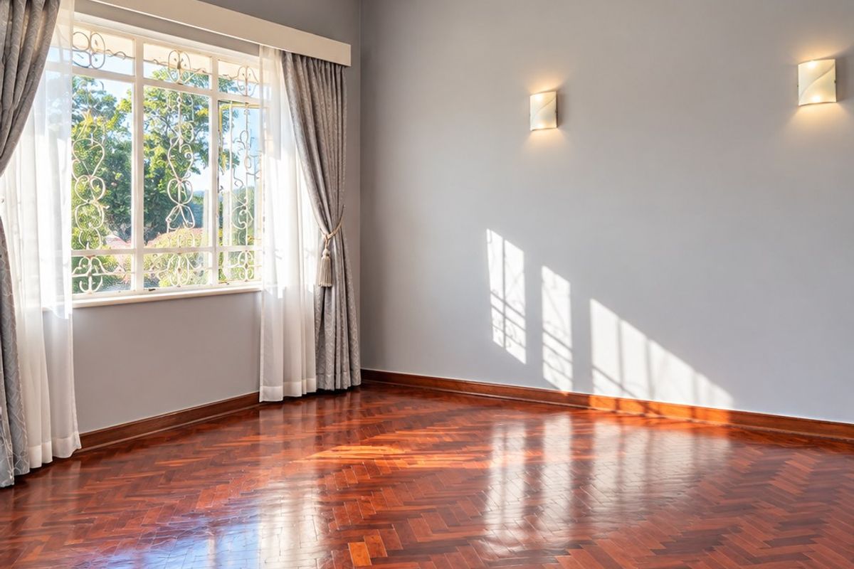 A bright, empty living room featuring polished herringbone parquet flooring, elegant wall sconces, a central chandelier, and large windows with decorative security bars.