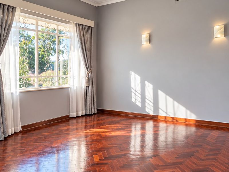 A bright, empty living room featuring polished herringbone parquet flooring, elegant wall sconces, a central chandelier, and large windows with decorative security bars.