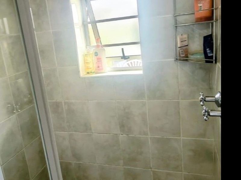A compact shower enclosure featuring tiled walls, a louver window, and a standard shower mixer, showing signs of wear and dated finishes.