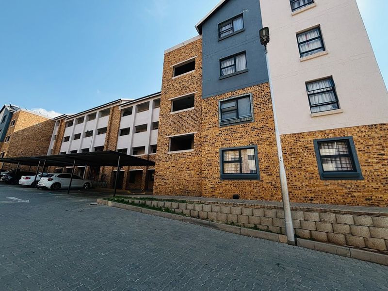 Exterior view of a multi-story apartment building featuring a brick facade, covered parking bays, and paved driveway access.