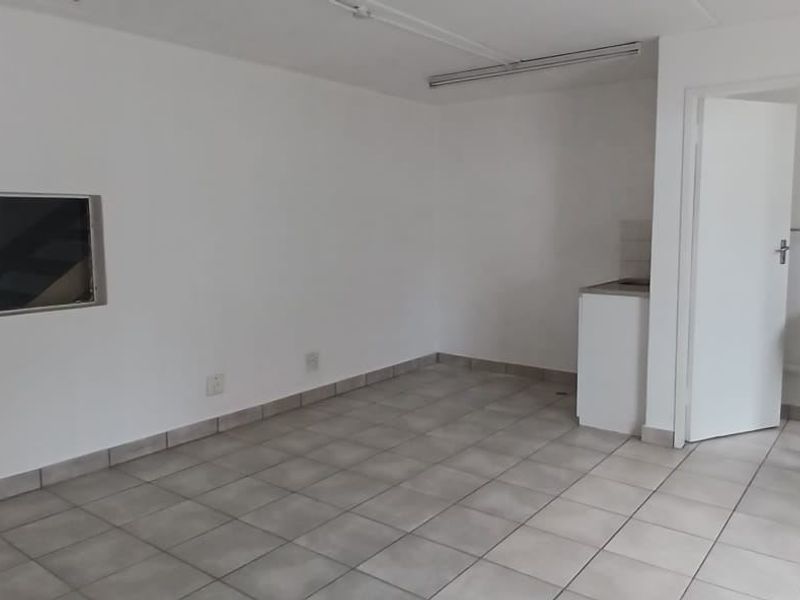 An open-plan industrial space featuring tiled flooring, a compact kitchenette unit, and direct access to an adjoining room.