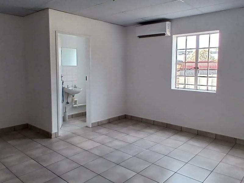 A clean, empty office space featuring tiled flooring, a drop ceiling, a wall-mounted air conditioner, and an attached private bathroom.