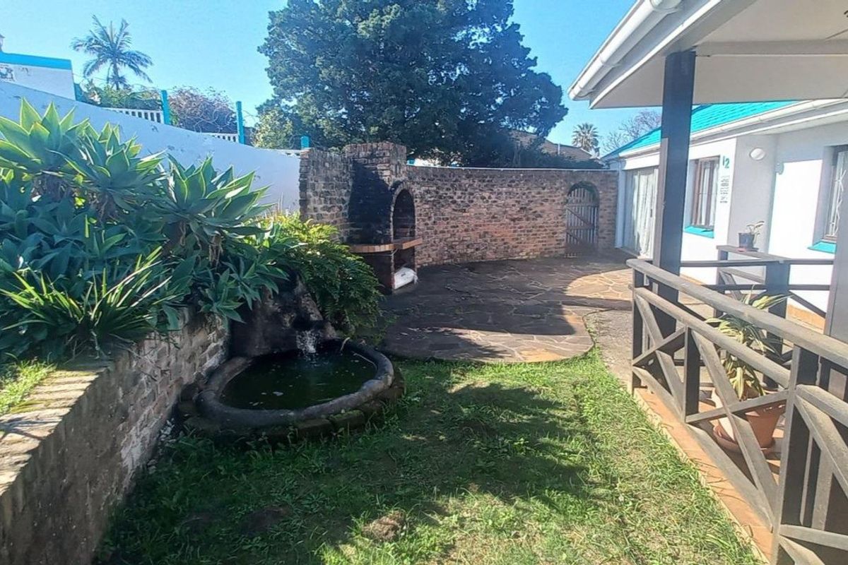 Rear garden area featuring a stone paved patio, built-in brick braai, water feature, and a covered veranda with timber railing.