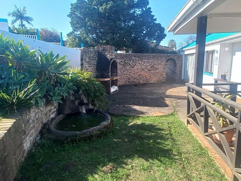 Rear garden area featuring a stone paved patio, built-in brick braai, water feature, and a covered veranda with timber railing.