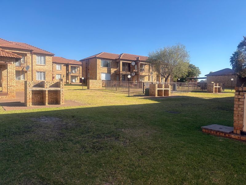 A view of the communal garden area featuring brick braai stands, well-maintained lawns, and the exterior facade of the apartment complex.