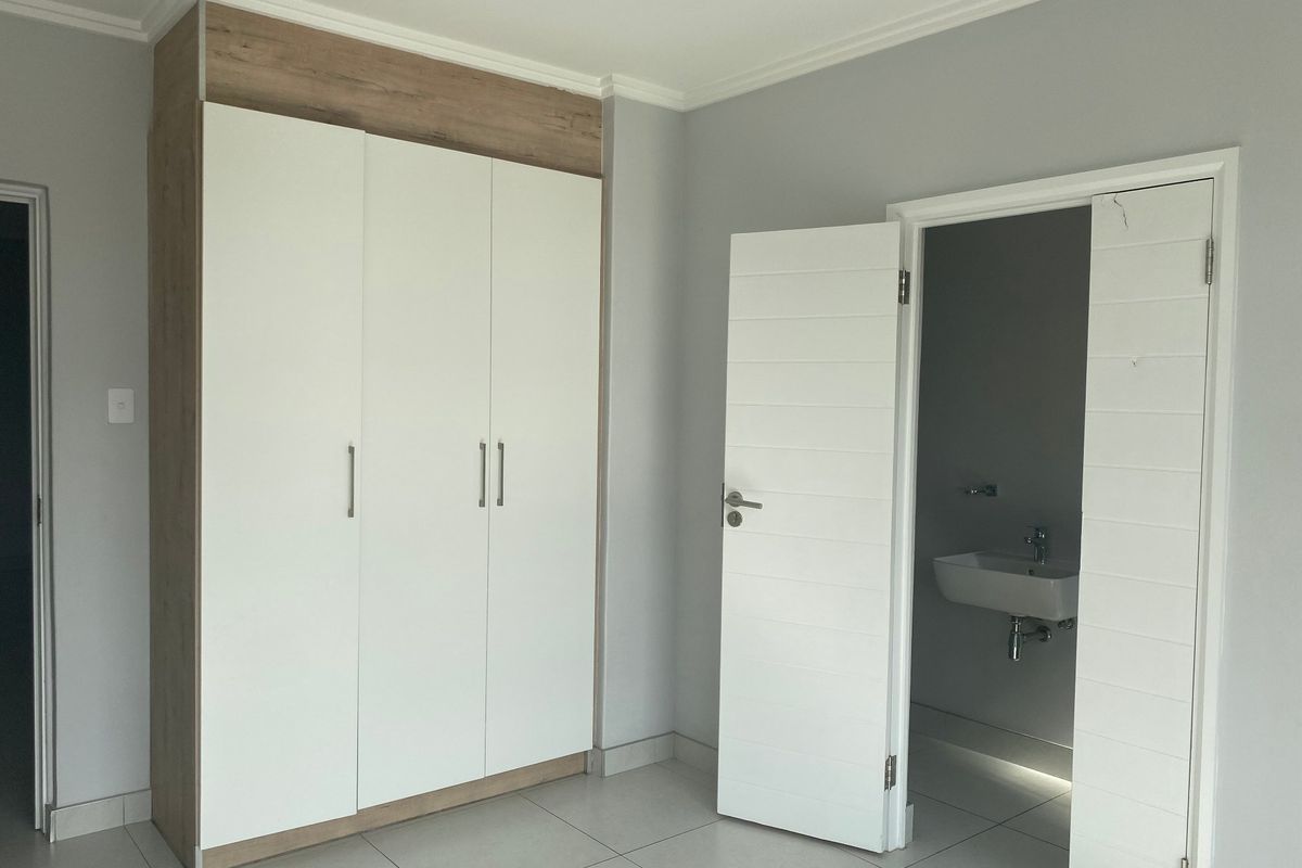 A clean, empty bedroom featuring modern built-in wardrobes with wood-look accents, neutral tiled flooring, and direct access to an en-suite bathroom.