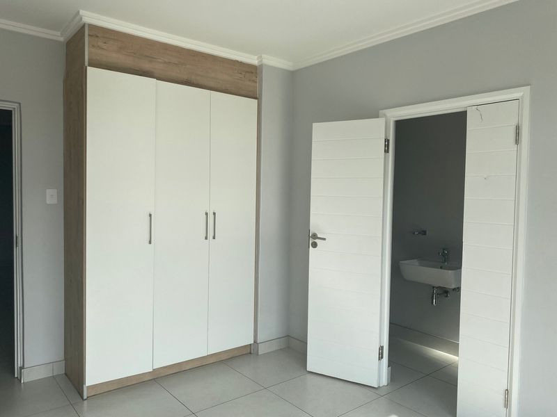 A clean, empty bedroom featuring modern built-in wardrobes with wood-look accents, neutral tiled flooring, and direct access to an en-suite bathroom.
