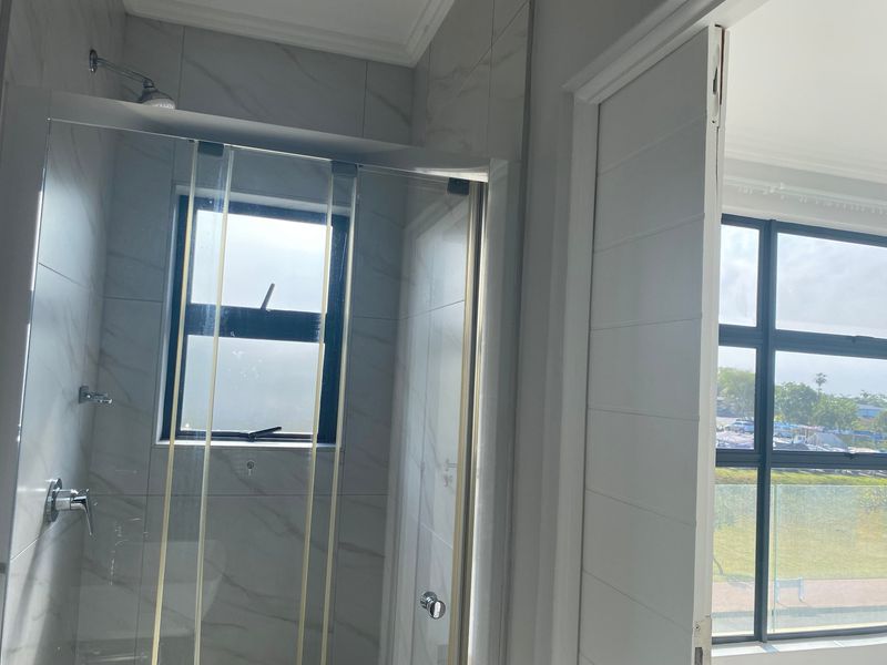 A modern bathroom featuring a glass-enclosed shower with marble-look wall tiles and large windows providing natural light.