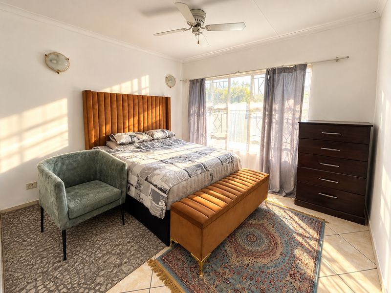 A well-lit bedroom featuring tiled flooring, a ceiling fan, wall sconces, and a security gate over the window.