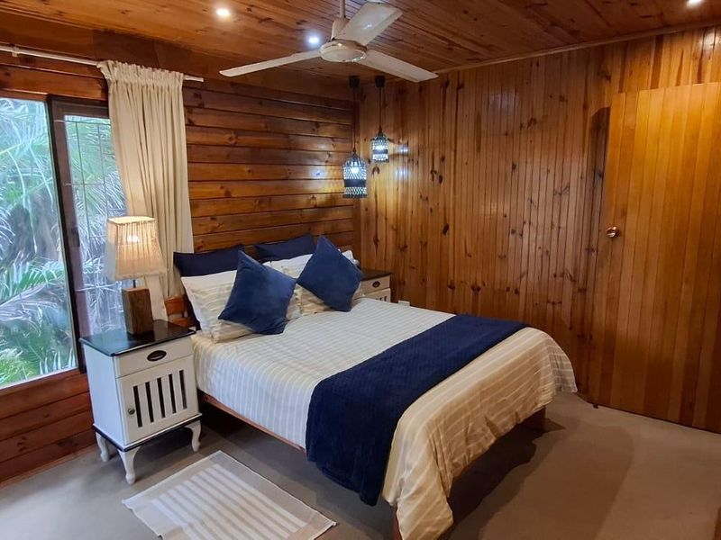 Cozy bedroom featuring warm timber wall paneling, a timber ceiling with a built-in fan, and large windows overlooking lush greenery.
