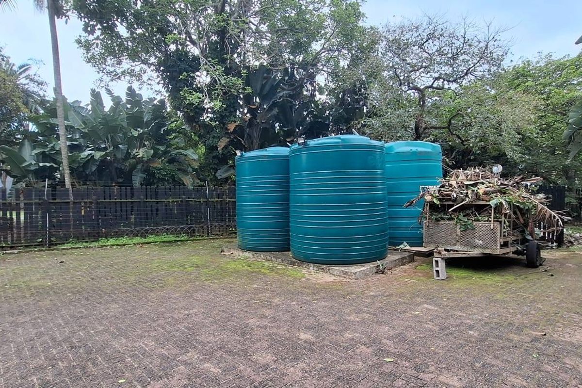 Rear exterior area featuring multiple water storage tanks, paved ground surface, and perimeter timber fencing surrounded by mature trees and garden vegetation.
