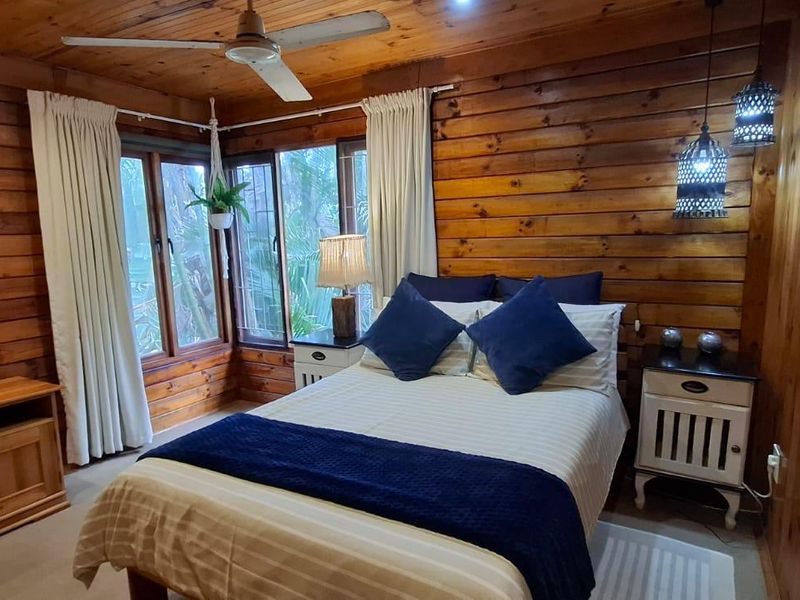 A cozy bedroom featuring warm timber wall paneling, a timber ceiling with a built-in fan, and large windows allowing natural light.